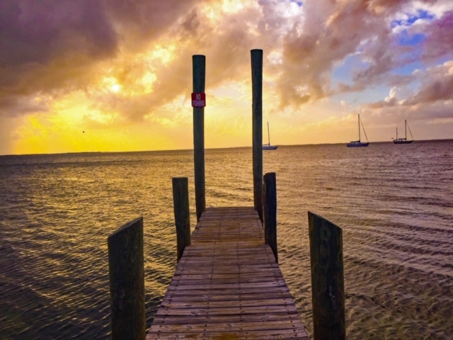 Wooden dock extending into calm ocean waters during a colorful sunset, with sailboats visible on the horizon and dramatic clouds in the sky