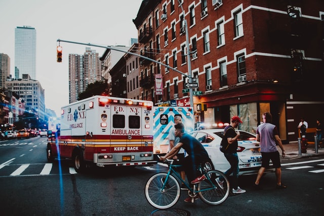 Ambulance with flashing lights stopped at a city intersection as pedestrians cross and a cyclist rides past, with buildings and traffic in the background