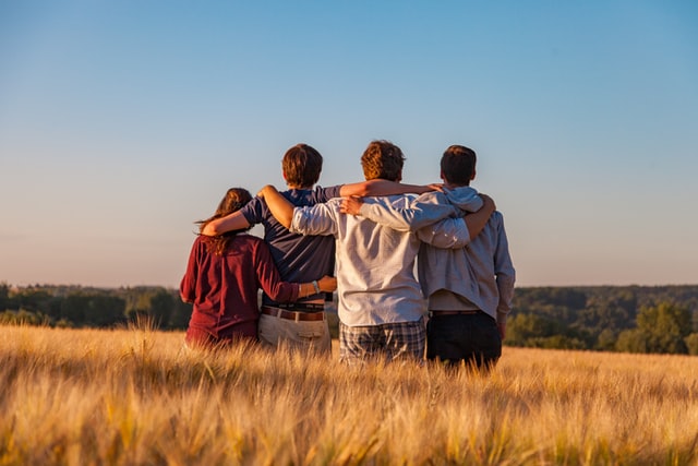 Group of four teenagers standing in a field with arms around each other, facing the horizon, symbolizing friendship, support, and connection